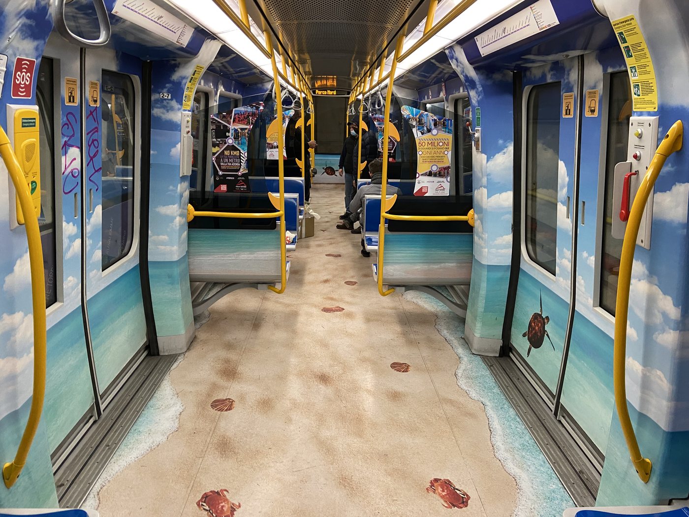 Interno di un treno M5 vestito a tema estate/mare — pareti con cielo e nuvole, pavimento sabbia con granchi e tartarughe